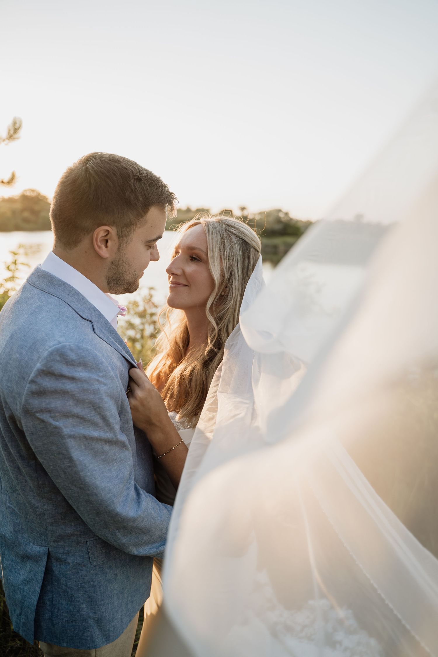Bride and groom share romantic golden hour moment outdoors with flowing veil and lake backdrop.