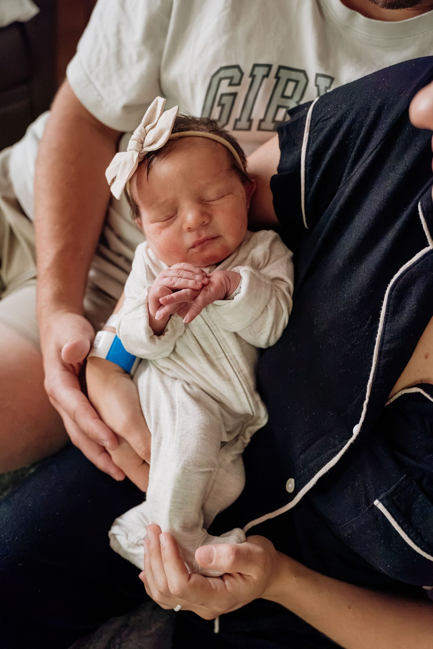 Newborn baby in white onesie with bow headband sleeping in parents arms at hospital.