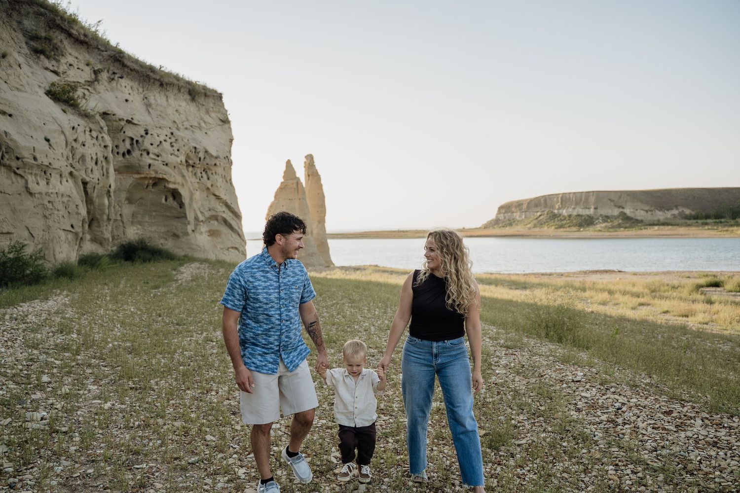 Young family of three walking hand-in-hand near dramatic sandstone cliffs and a lake at sunset.