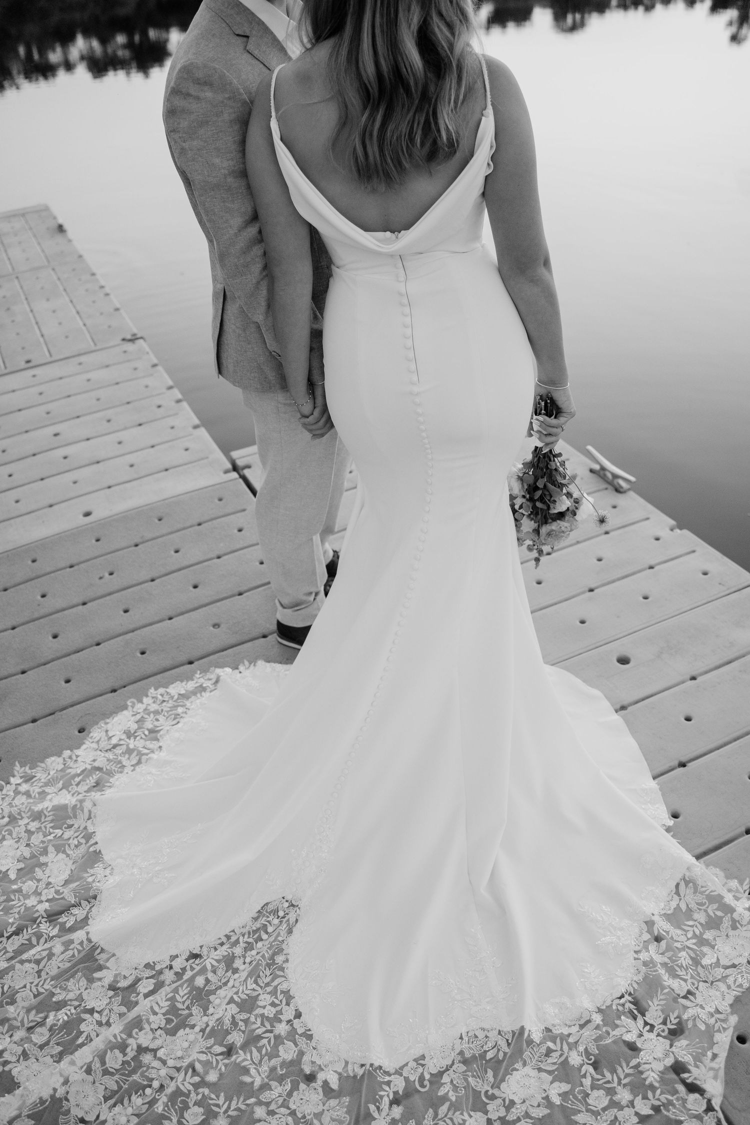 Black and white photo of bride's elegant mermaid wedding gown train on a wooden dock by a lake.