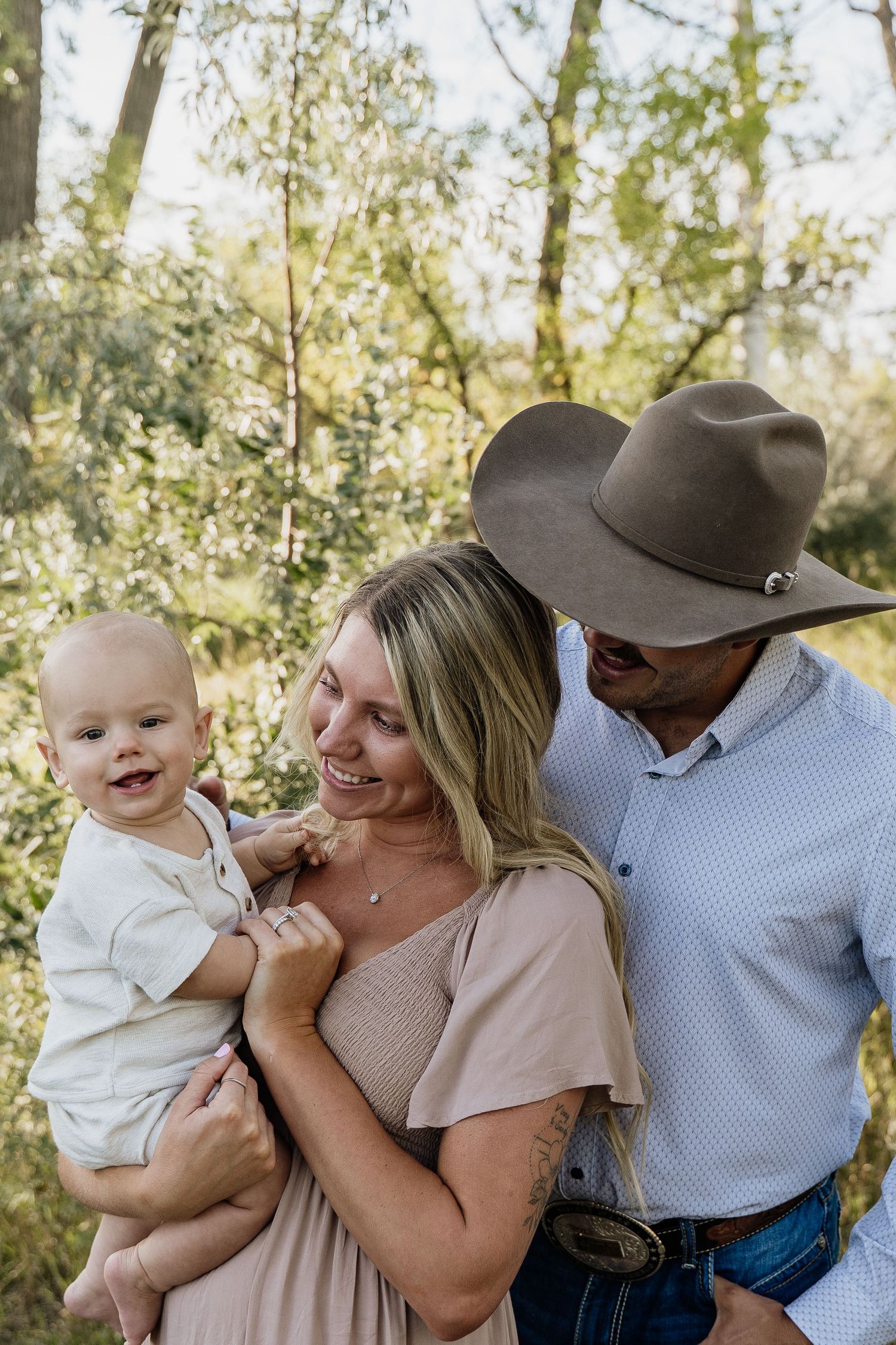 Happy young family with baby outdoors, mother holding infant while father in cowboy hat smiles in green nature setting.