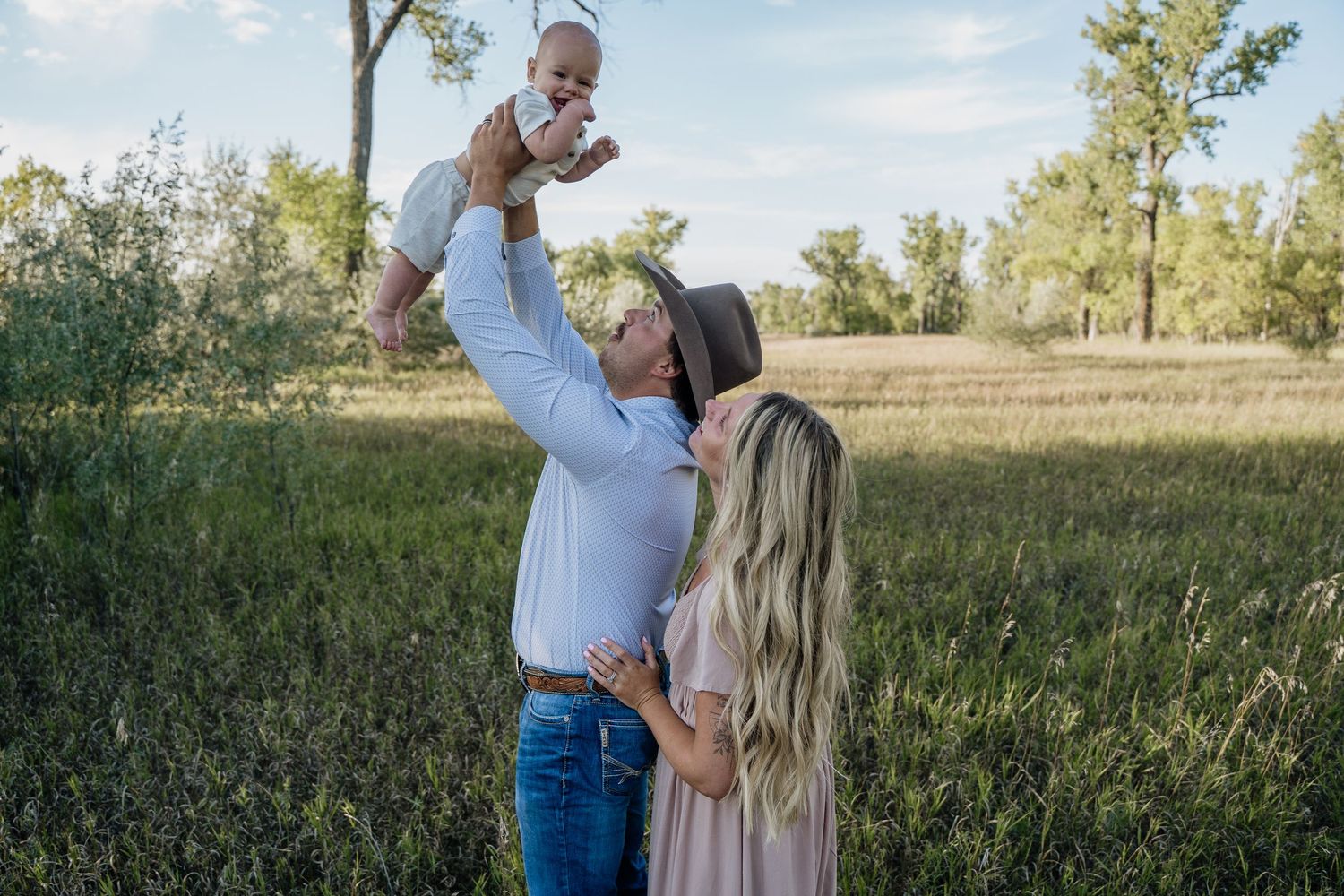 A happy family outdoors, father lifting baby in air while mother watches in a lush green field.
