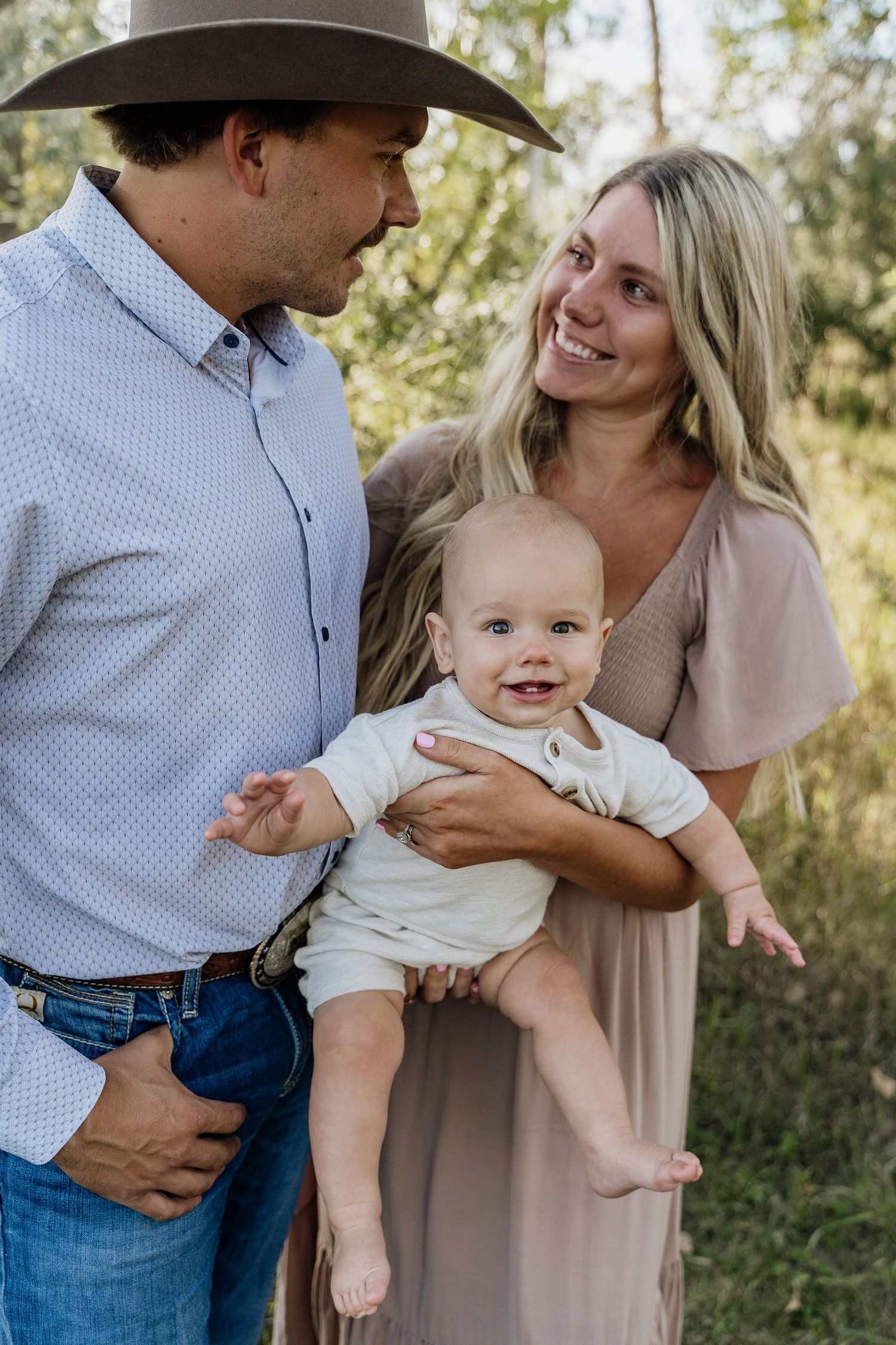 A smiling family with a cowboy hat-wearing father and blonde mother holding their happy baby outdoors.