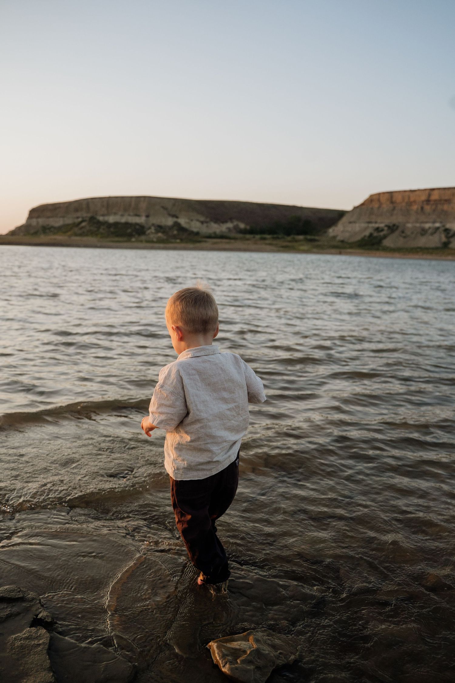 Curious toddler in a white shirt wades alone in shallow water with majestic cliffs in the background.