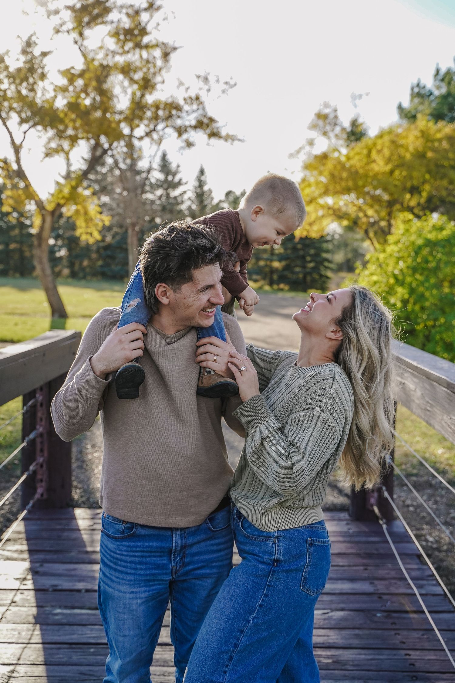 Happy family with baby on fathers shoulders laughing on a wooden bridge outdoors.