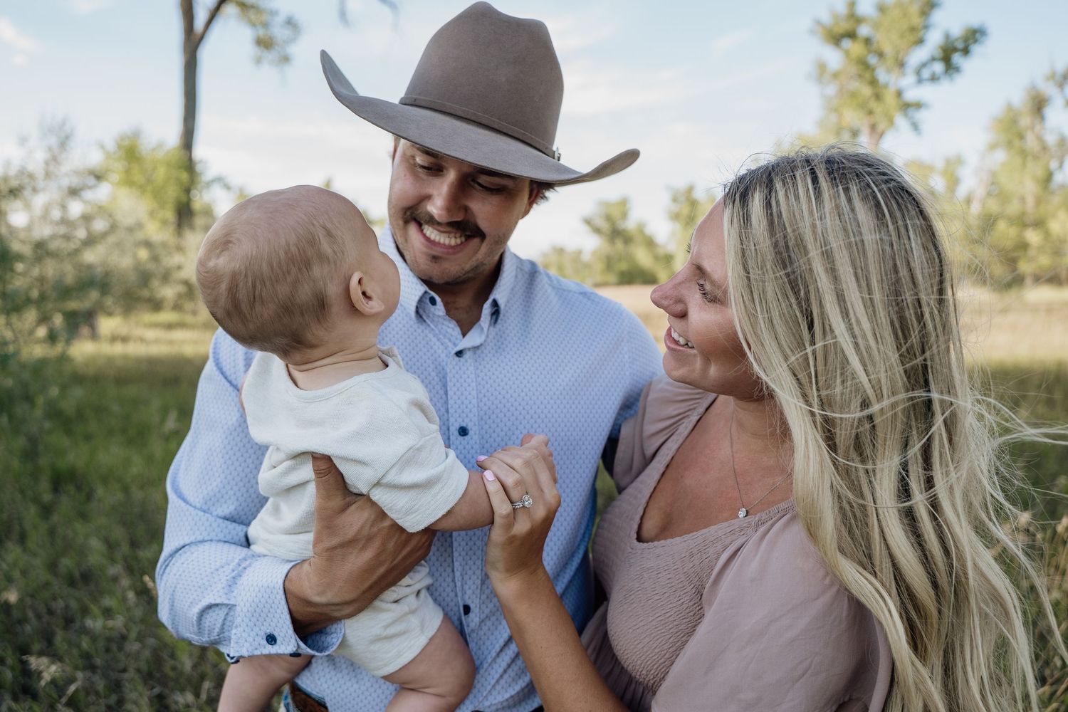 Smiling father in cowboy hat holds baby while mother looks on lovingly, outdoor family portrait in natural setting.