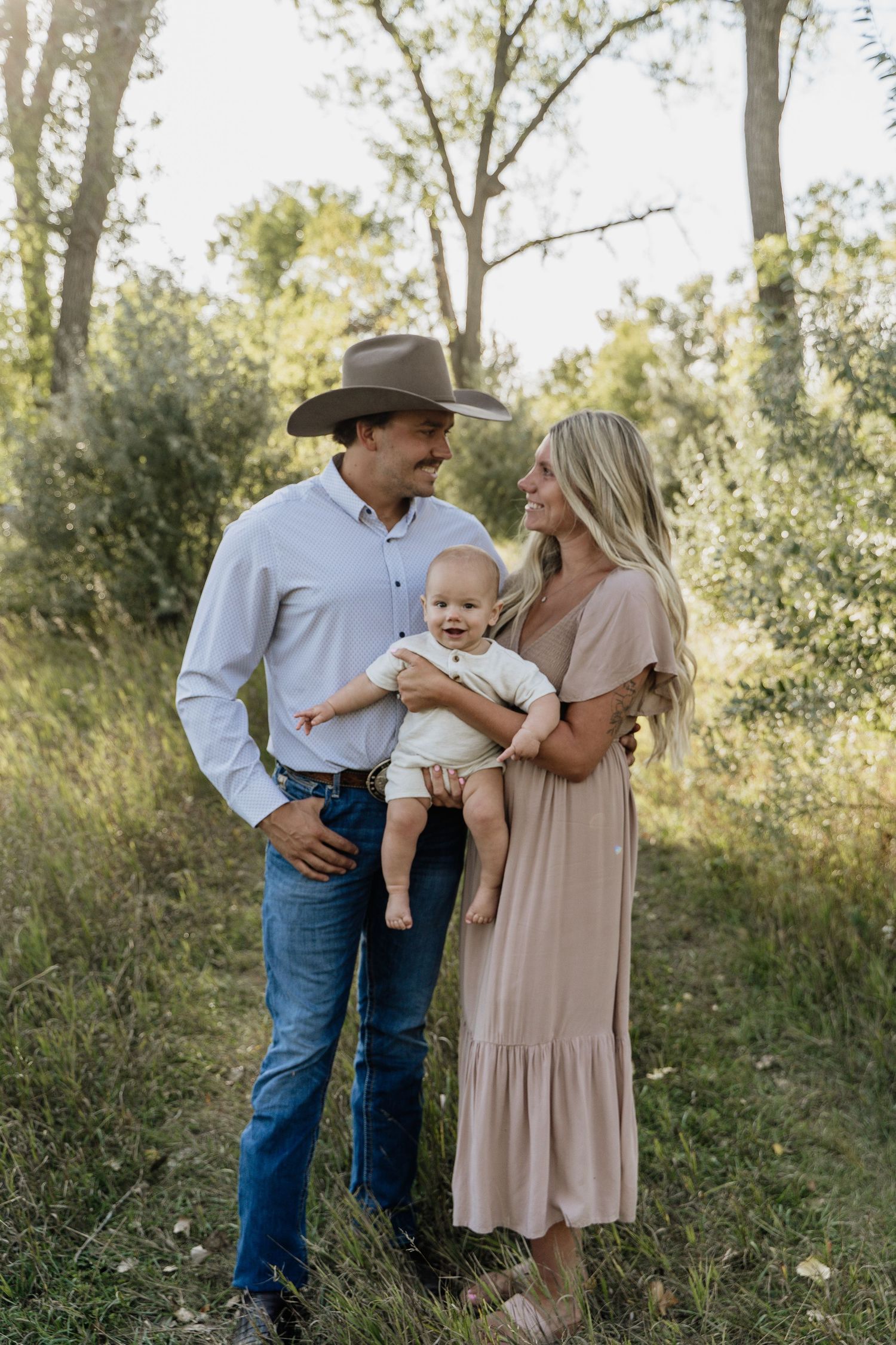 Young family with baby poses outdoors, father in cowboy hat and jeans, mother in blush maxi dress, green trees behind.