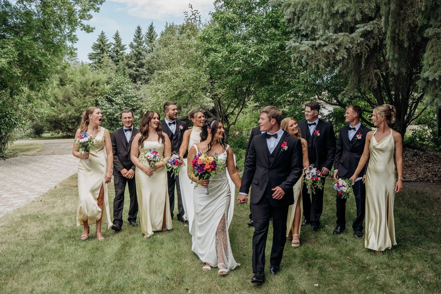 Wedding party walking together outdoors in cream dresses and black suits on a sunny summer day.