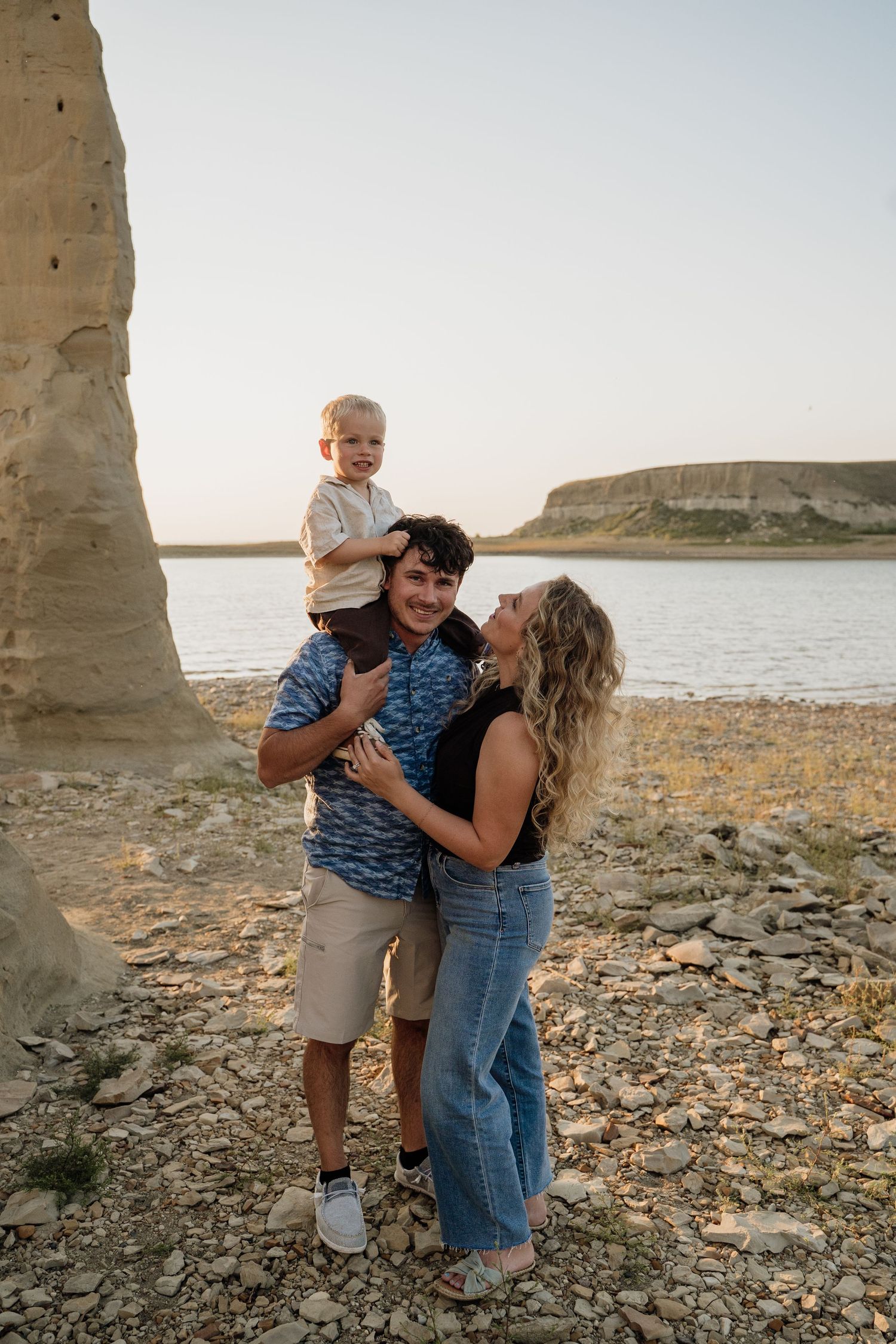 Happy family of three poses near a rocky shoreline with dramatic cliffs and calm water at sunset.