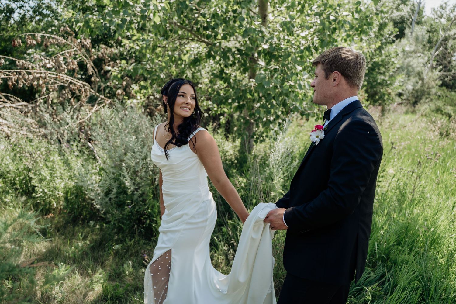 Bride in white gown smiles at groom who holds her train in a lush green outdoor setting.