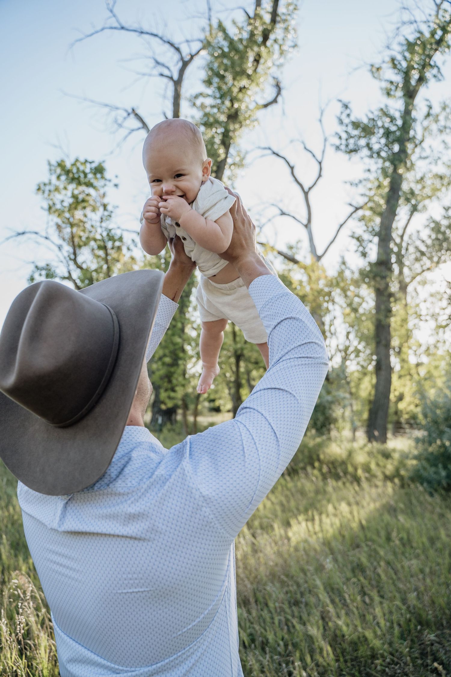 Father in cowboy hat lifting baby up in the air in a lush outdoor setting.