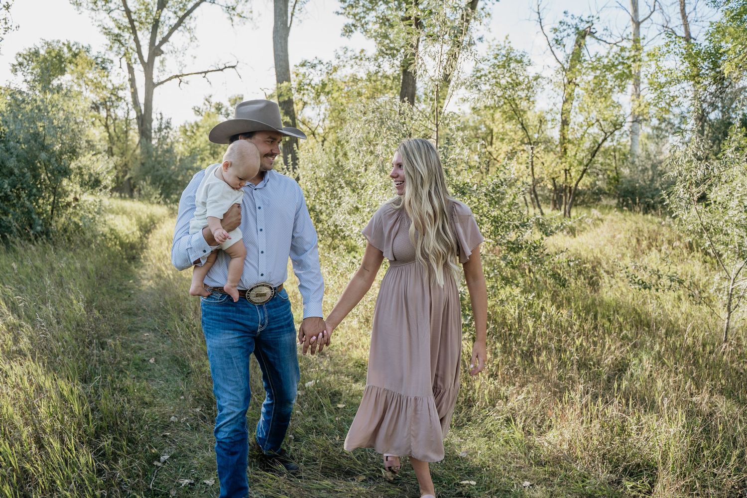 A couple in rustic attire walks hand-in-hand through a meadow path, man holding a baby, woman in a flowing mauve dress.