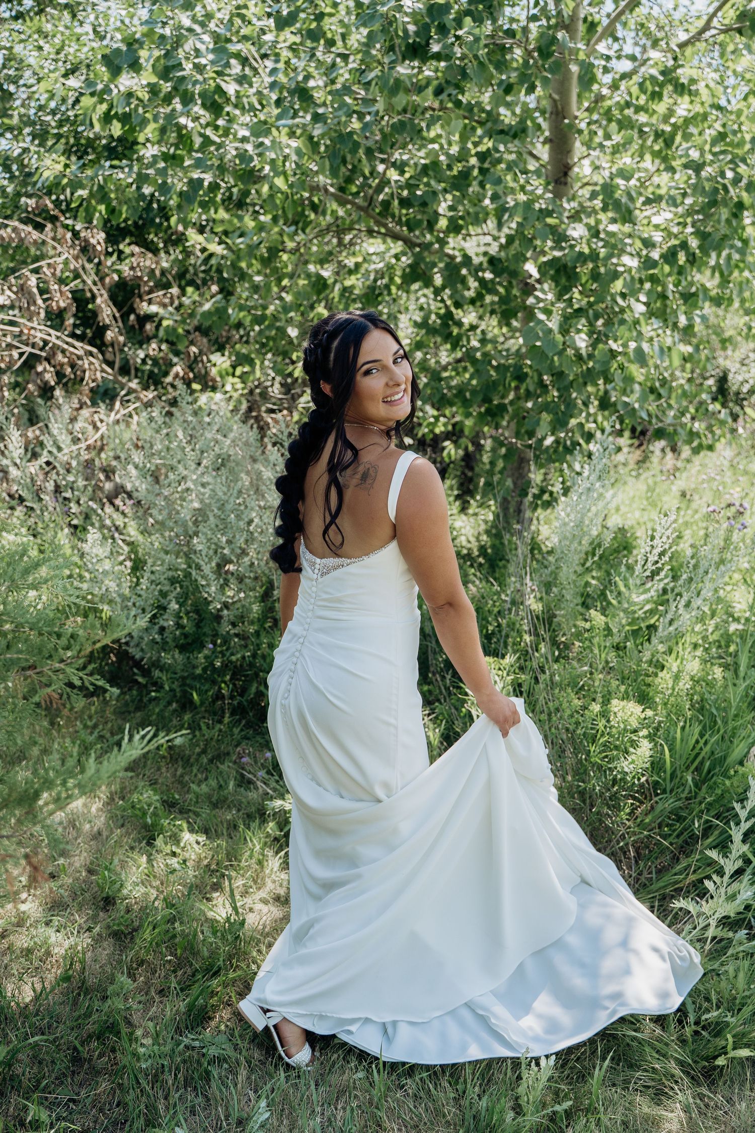 Smiling bride in a white satin wedding gown holding her train while standing in a lush green garden.