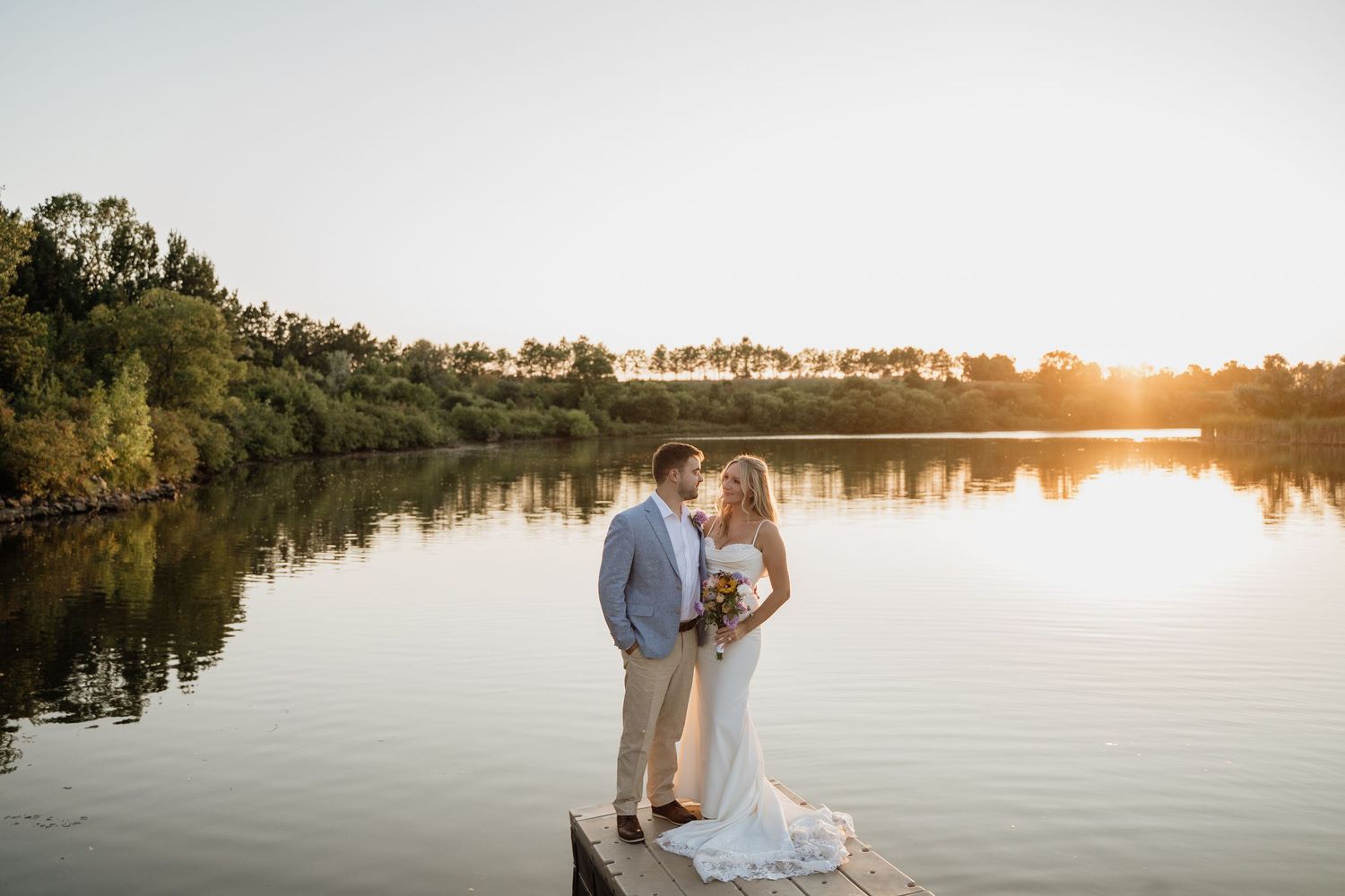 Bride and groom kissing on a dock with a stunning lake and golden sunset reflection behind them.