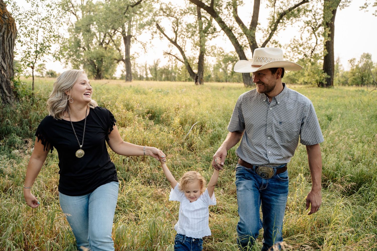 Happy family with cowboy dad swinging toddler between parents in sunny green field.