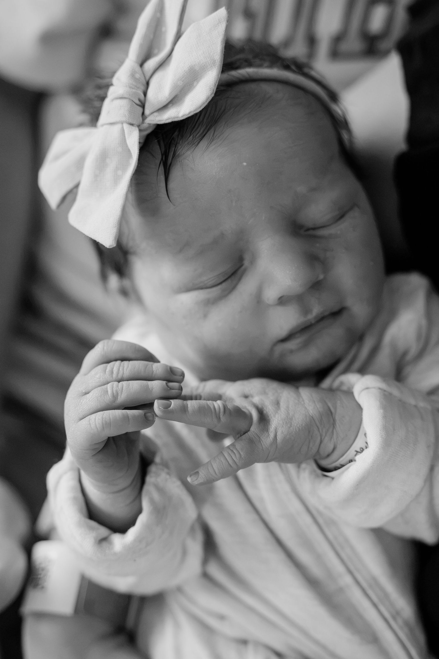 Black and white close-up of newborn baby girl with bow headband grasping adult finger gently.