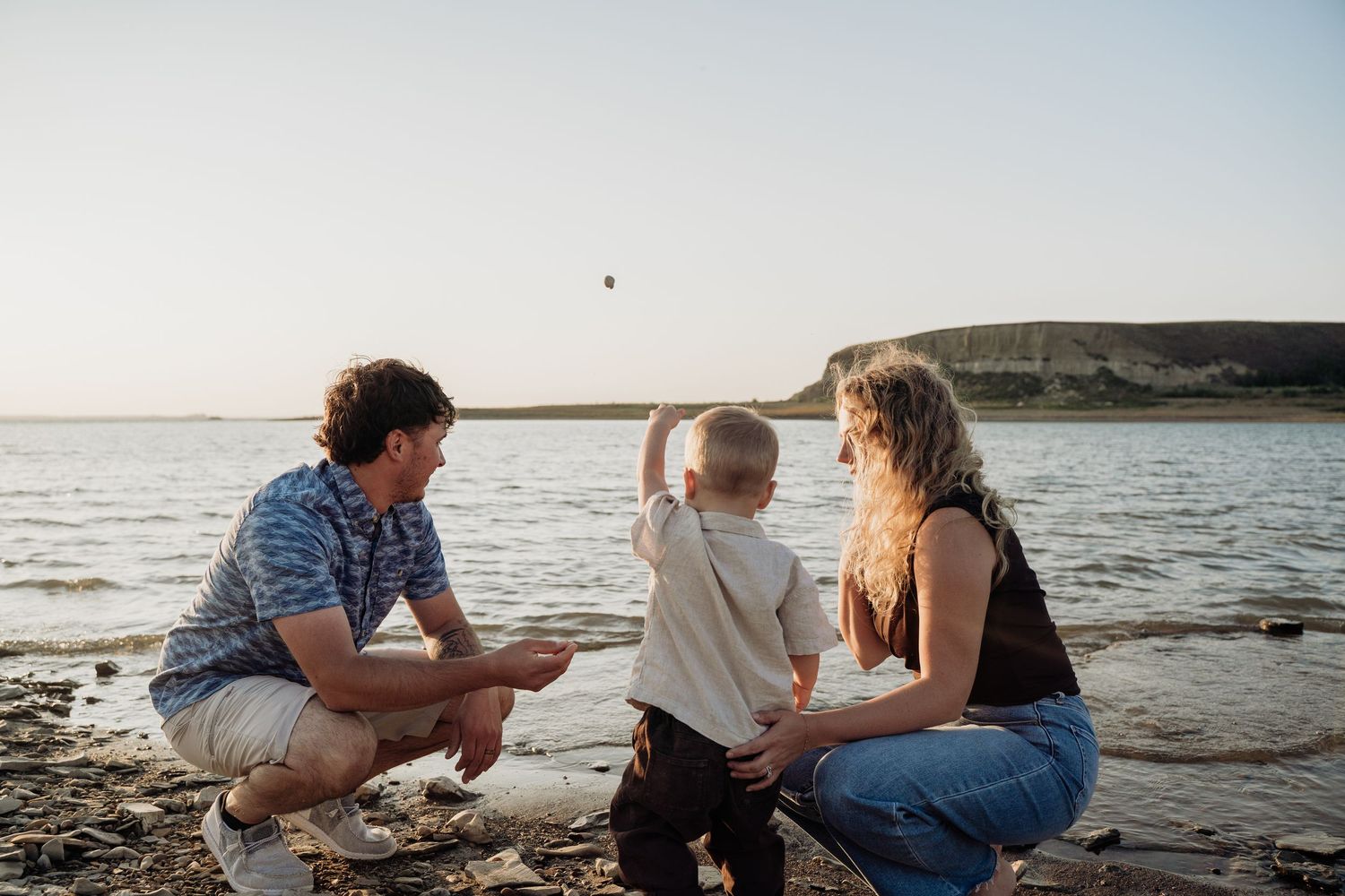 Parents crouching with toddler on rocky lake shore as child points toward the calm water.