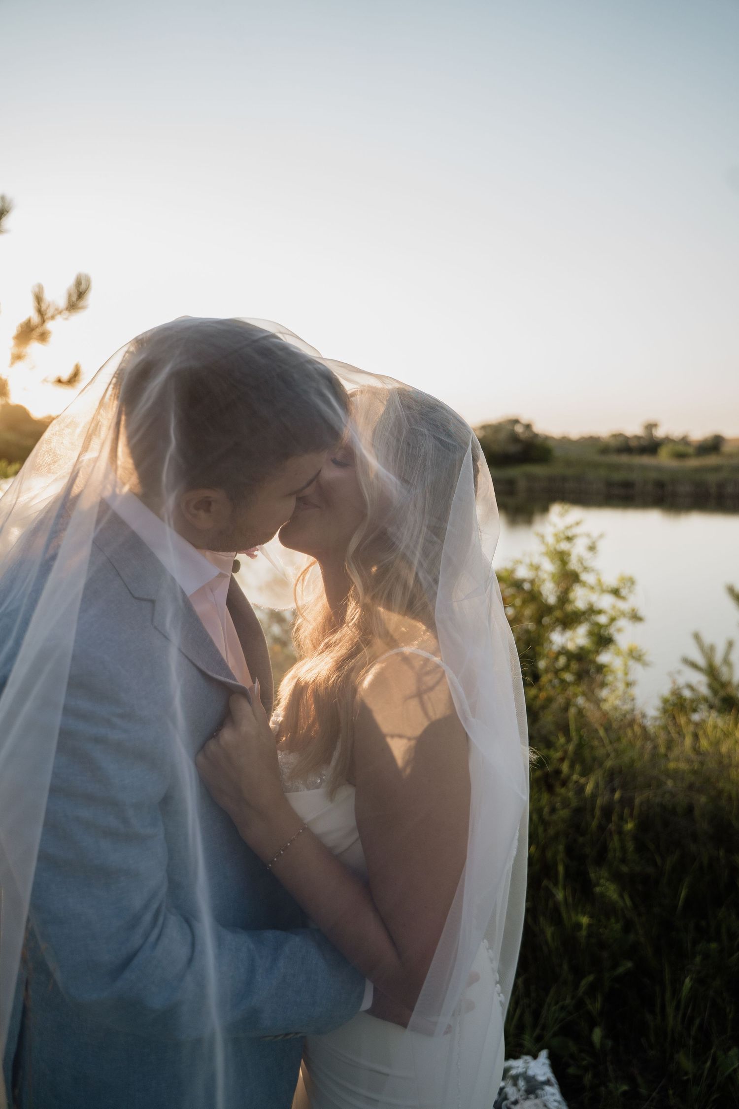 Bride and groom sharing an intimate moment under a veil near a serene lake at sunset.