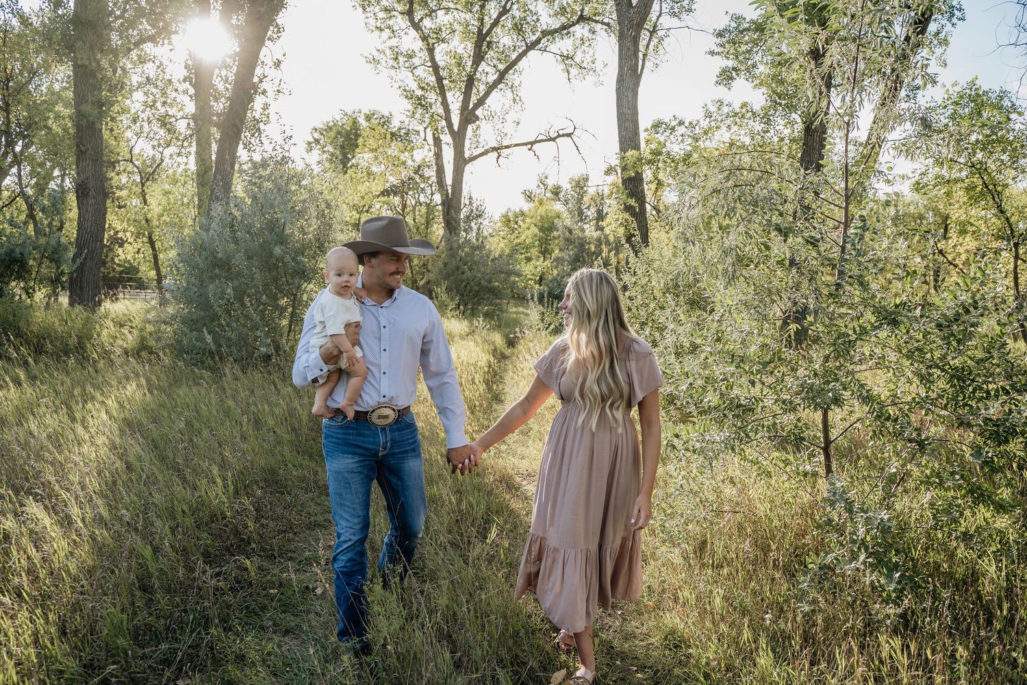 A couple in western attire walks hand-in-hand through a sunlit forest path, the man carrying their young baby.
