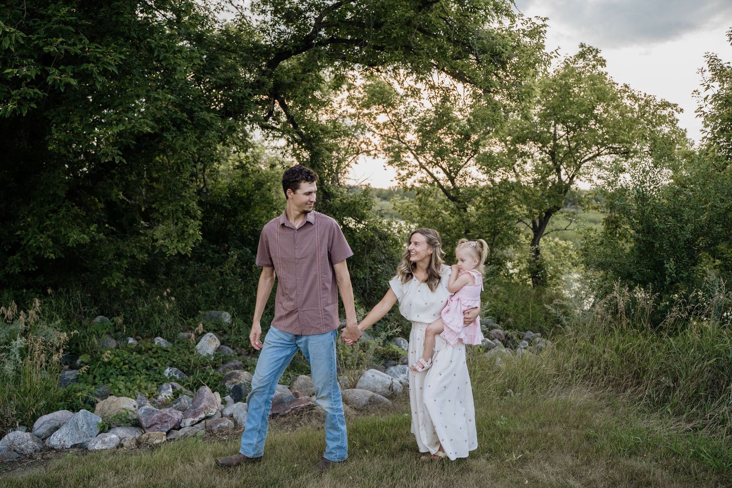 Young couple walking hand-in-hand through a green landscape, with the woman carrying a baby at golden hour.