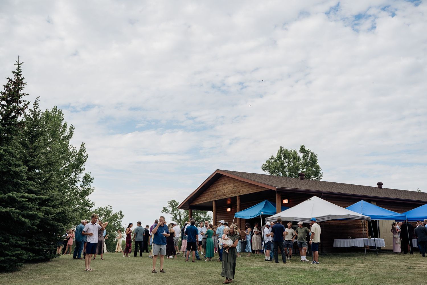 Outdoor wedding reception with guests mingling on a green lawn beside a rustic wooden venue under a partly cloudy sky.