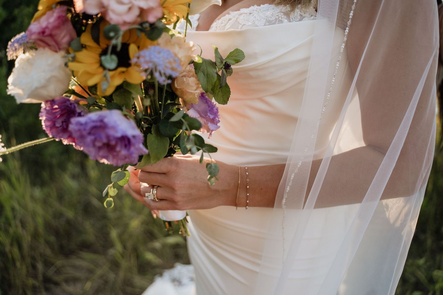 Bride holding colorful wedding bouquet with purple and yellow flowers wearing lace gown and veil.