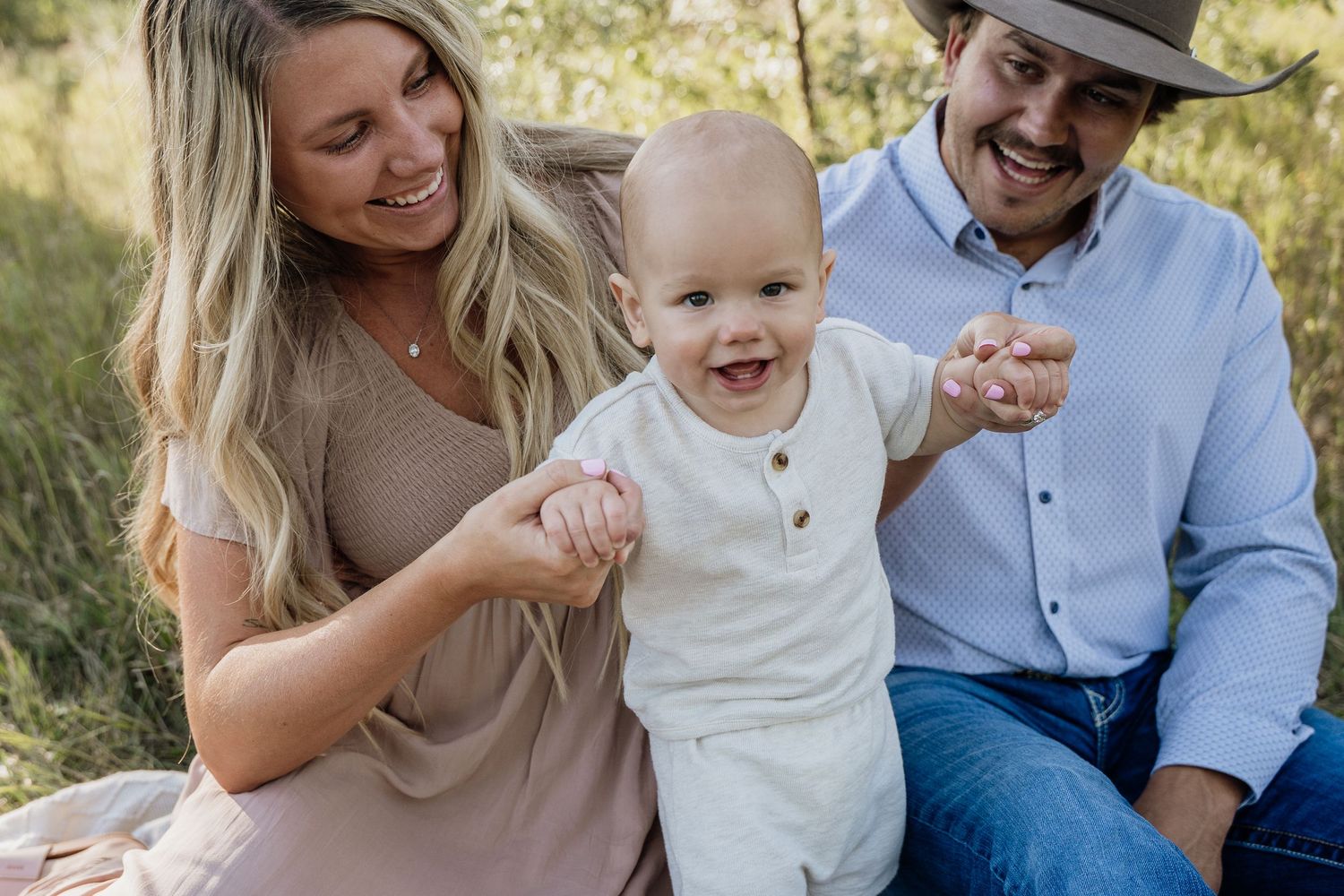 Happy family with smiling baby boy held by blonde mother and father in cowboy hat outdoors in nature.