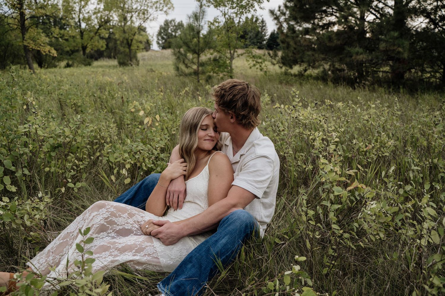 Couple sitting together in a lush green meadow, man kissing woman on the head during outdoor engagement session.