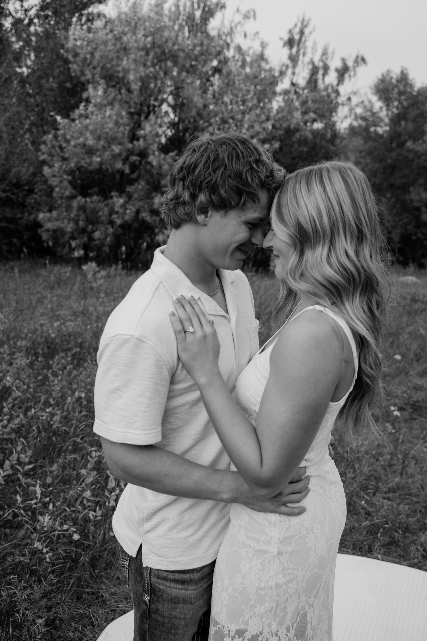 Black and white photo of a couple embracing and smiling at each other in a lush outdoor field setting.