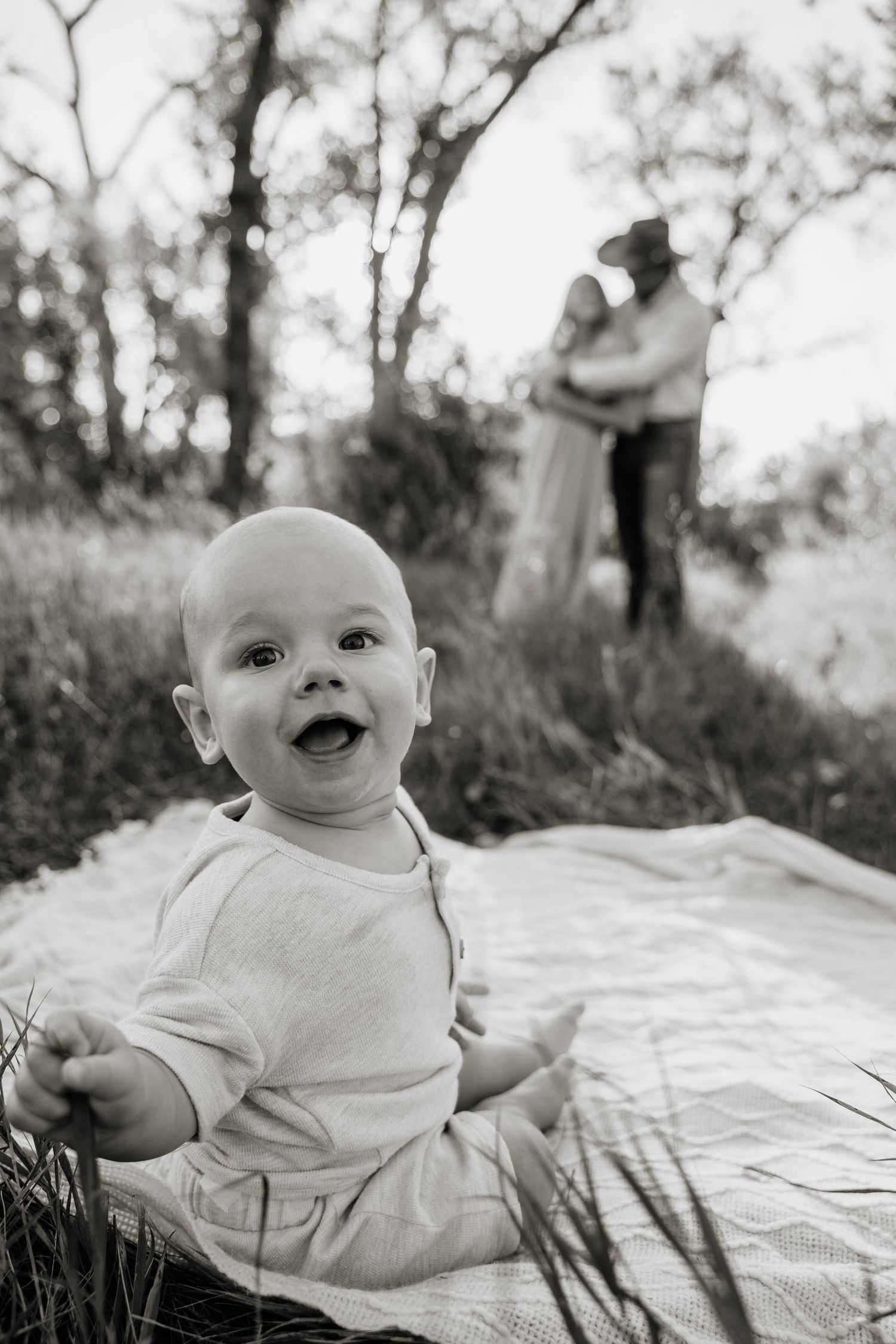 Black and white photo of a smiling baby sitting on a blanket outdoors with parents embracing in the background.
