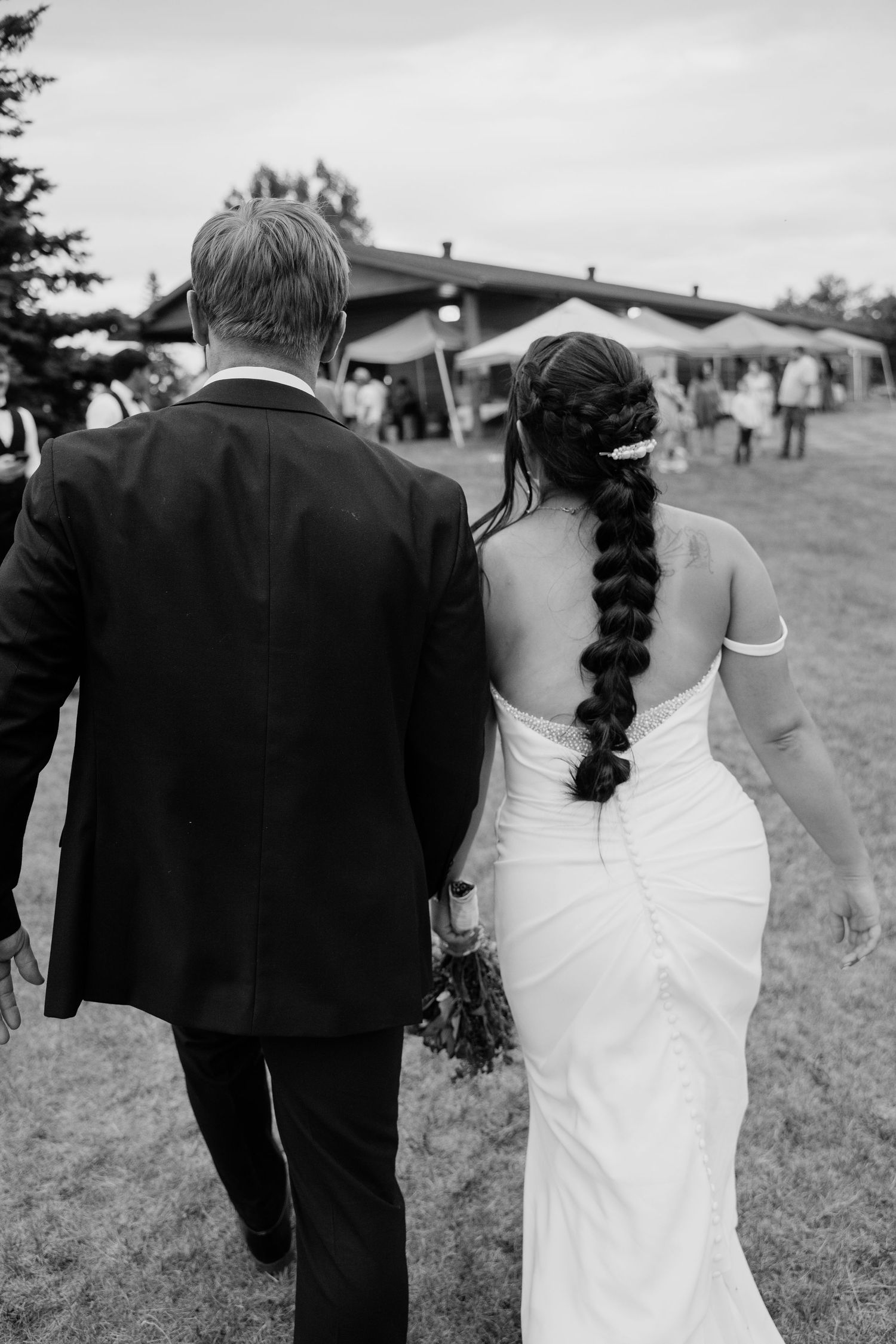 Black and white photo of bride with long braided hair and groom walking hand in hand toward wedding reception tent.