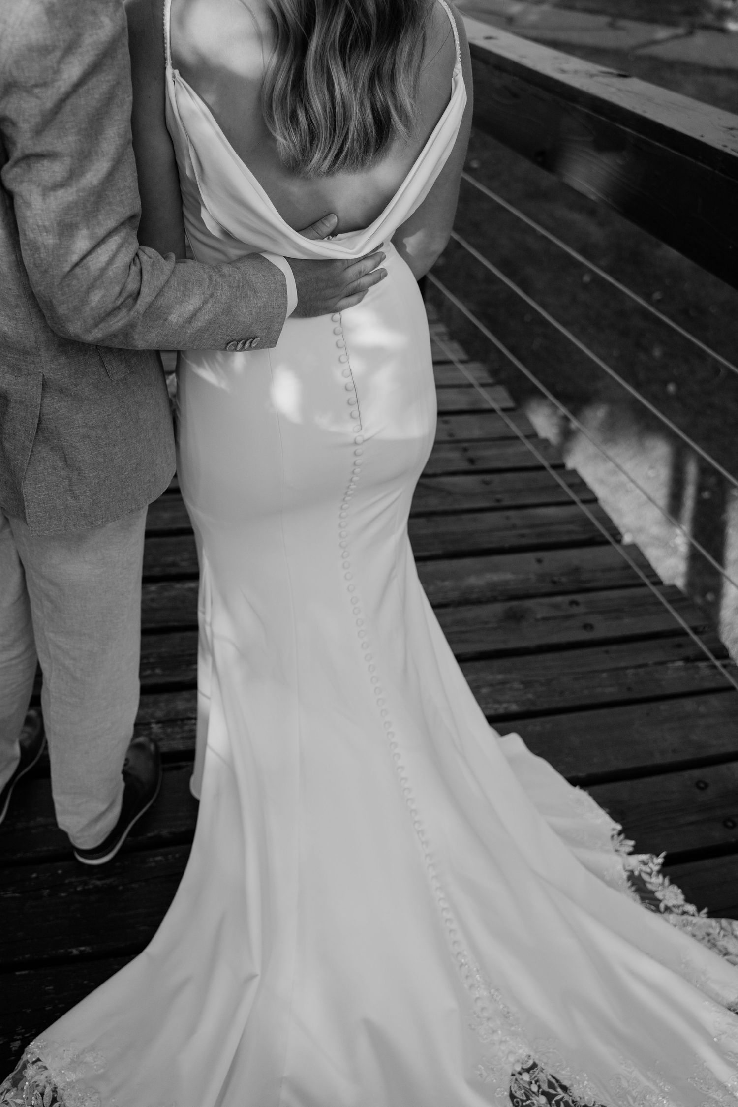 Black and white photo of bride and groom embracing on wooden dock showing elegant gown train.