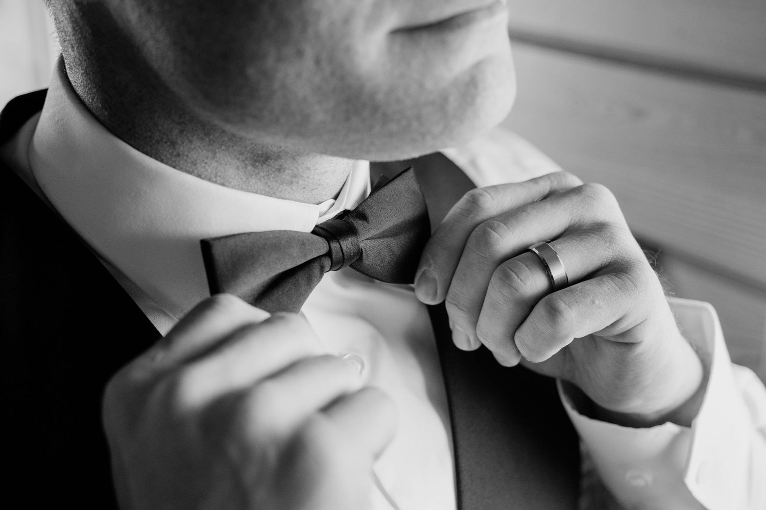 Black and white close-up of groom adjusting his bow tie while wearing a suit and ring.