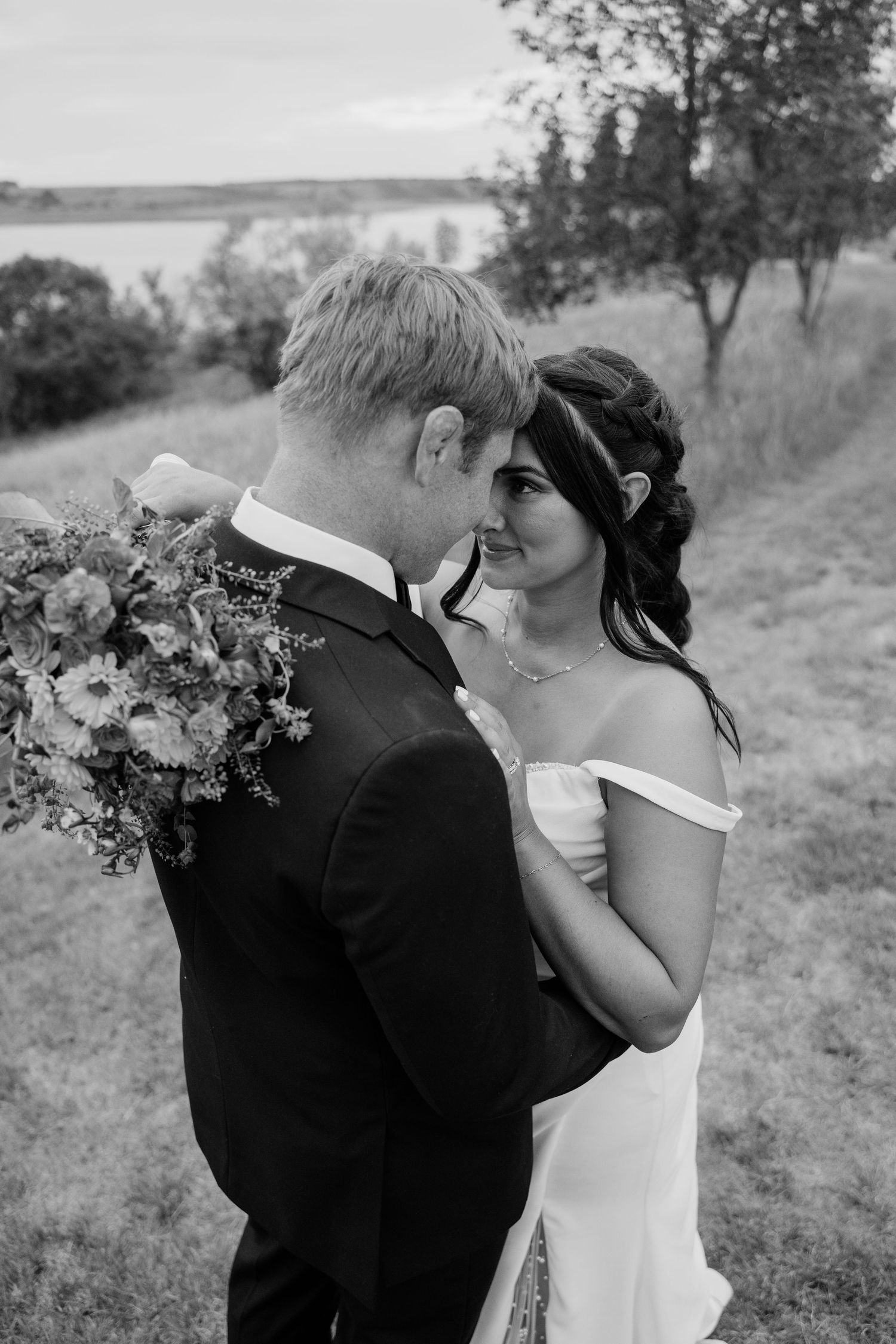 Black and white photo of bride and groom sharing a romantic moment near a lakeside meadow.