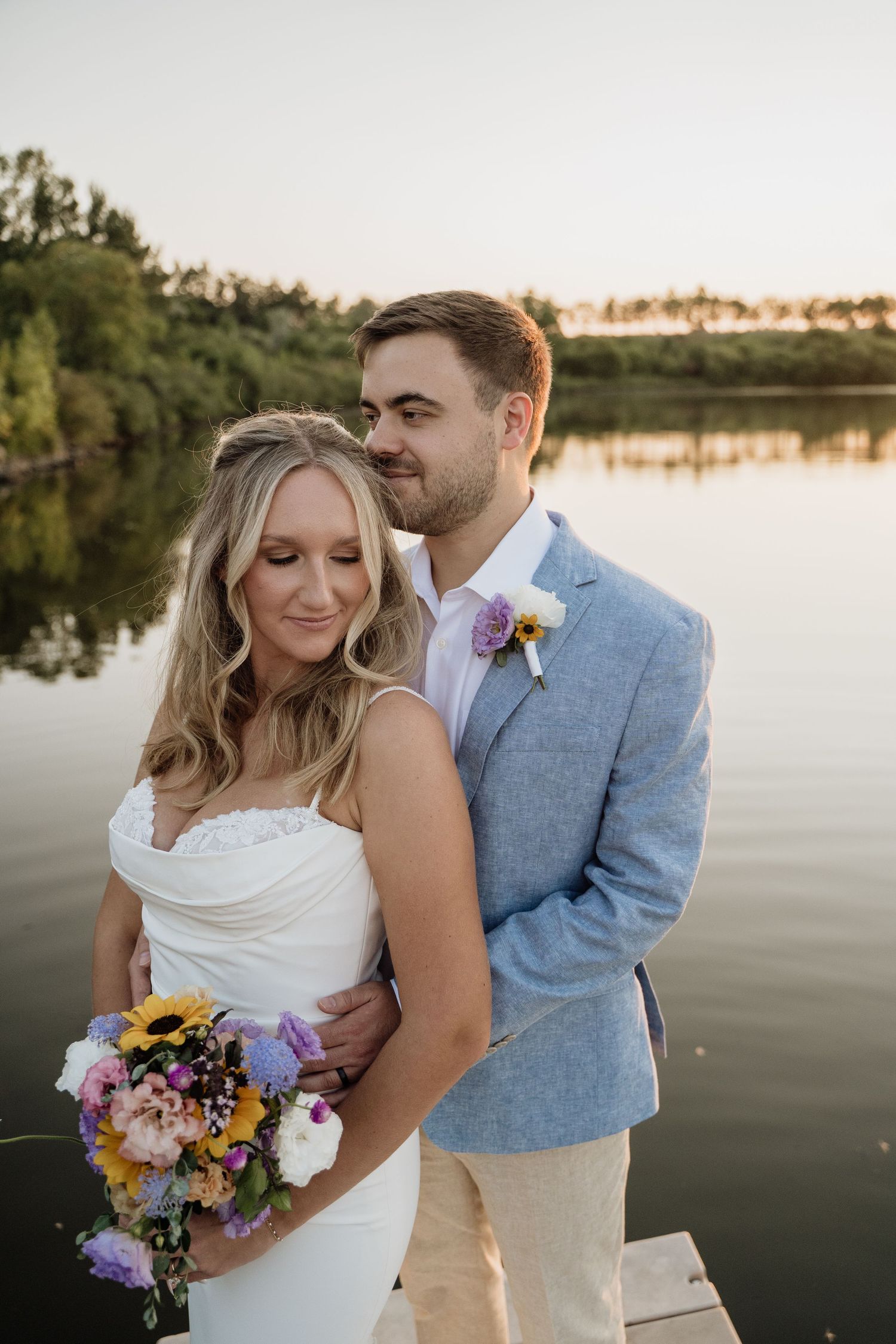 Bride and groom embracing on a dock at golden hour with a calm lake reflection behind them.