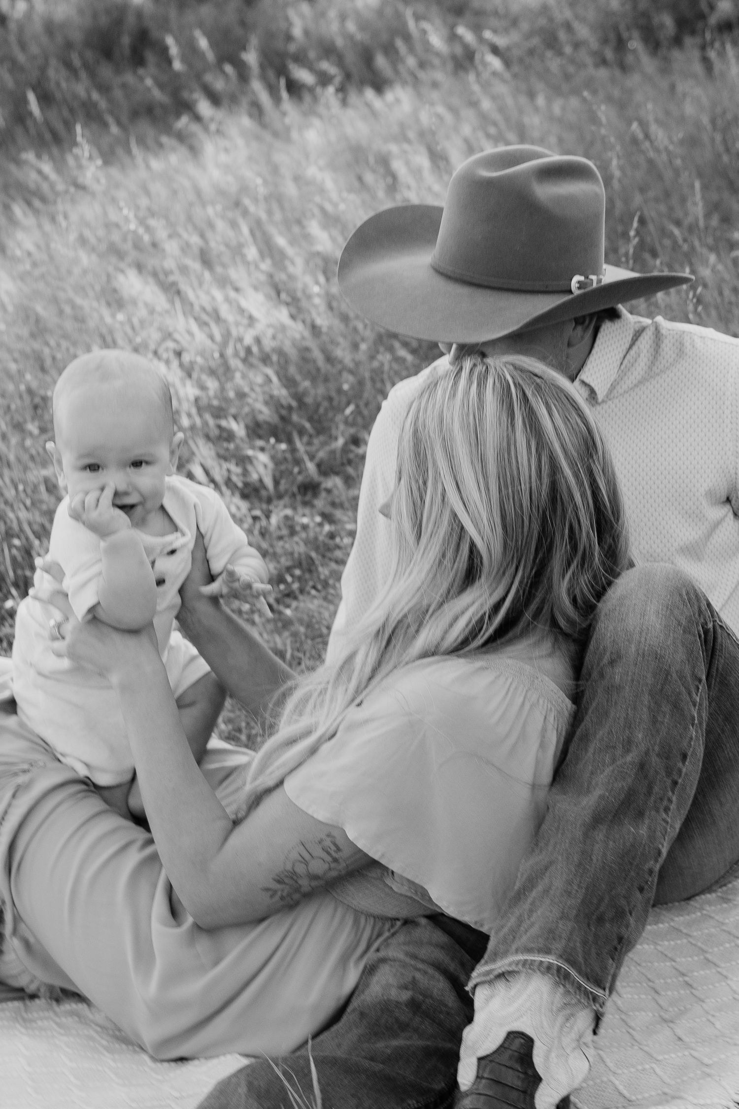Black and white photo of a couple sitting in a field with their baby, man wearing a cowboy hat, woman holding the infant.