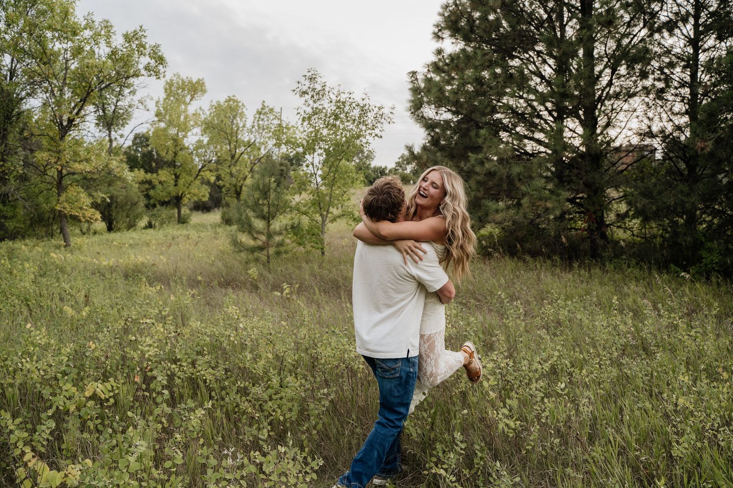 Couple embracing and laughing in a lush green meadow surrounded by trees during an outdoor engagement photo session.