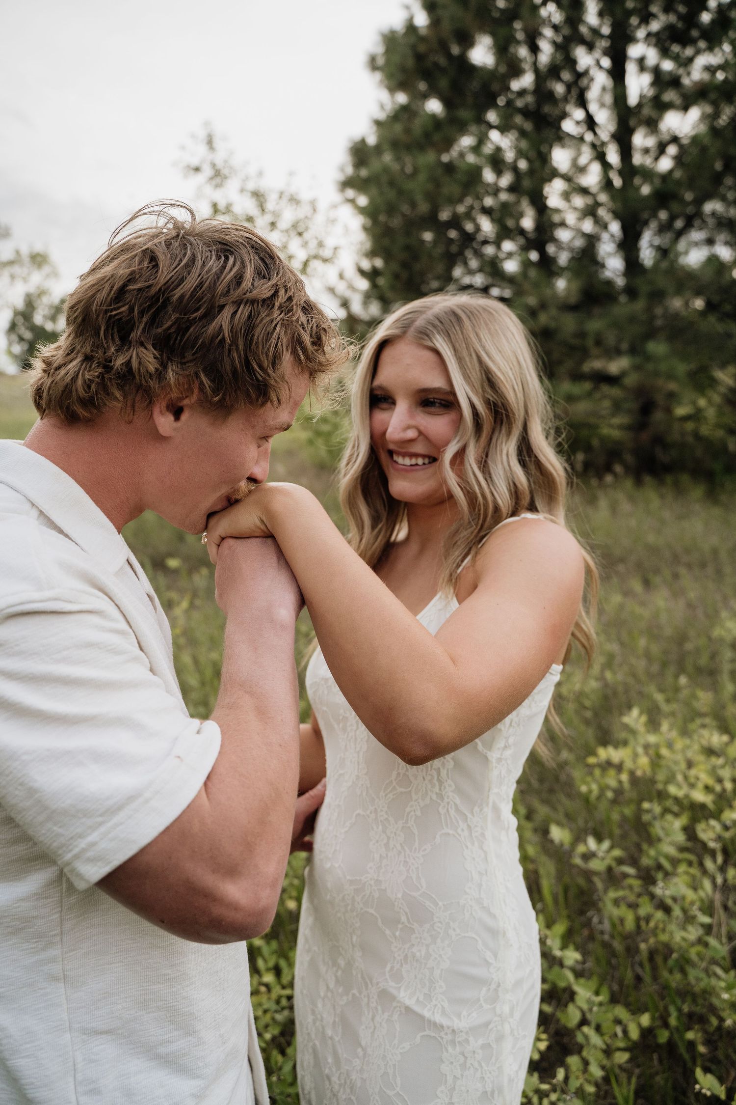 Man kissing woman's hand while she smiles joyfully, standing in lush green field, engagement photo.