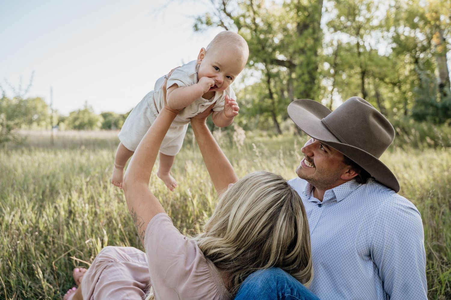 A couple sits in a green meadow, lifting their smiling baby in the air during a family photo session.