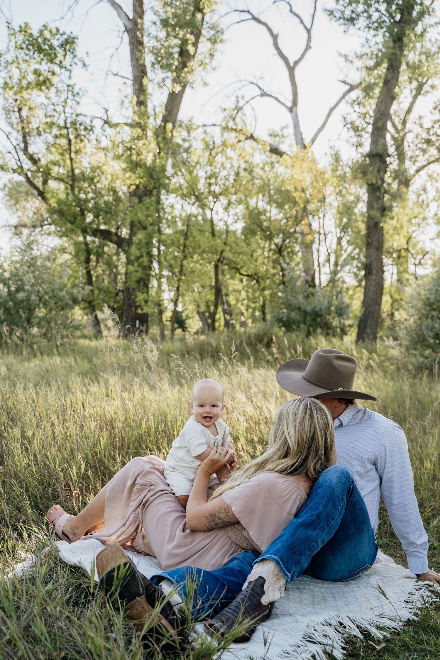 Young family sitting on blanket in green meadow with trees, parents enjoying time with happy baby.