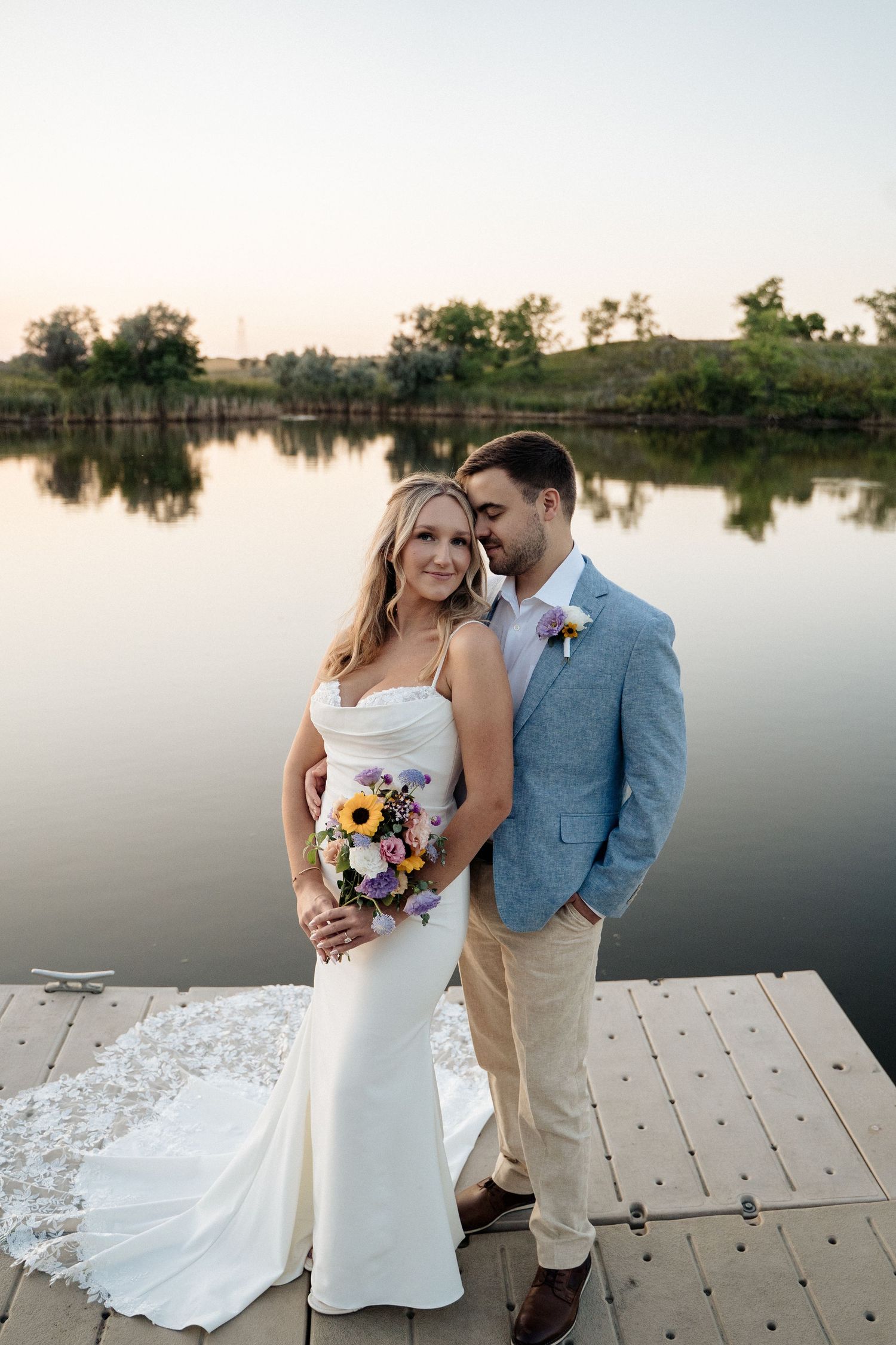 Bride and groom pose on a lakeside dock at sunset, bride holding colorful bouquet in elegant white gown.