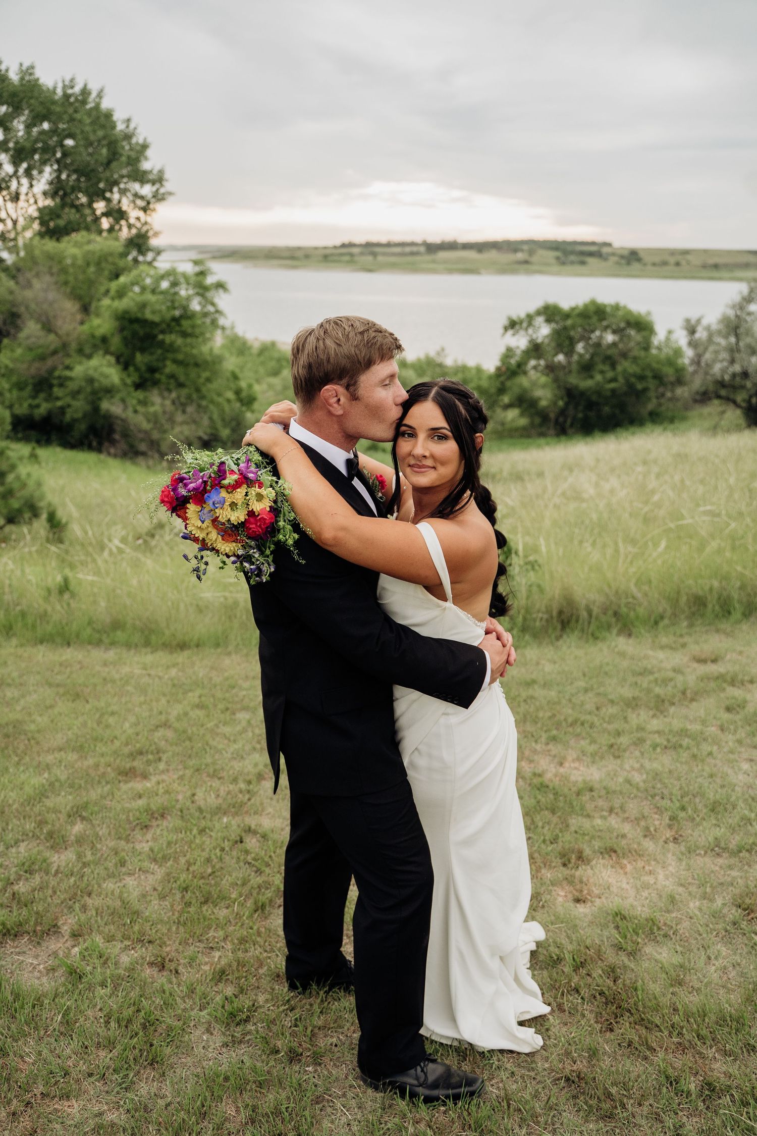 Groom kissing bride on the cheek as she smiles holding a colorful bouquet by a lake.