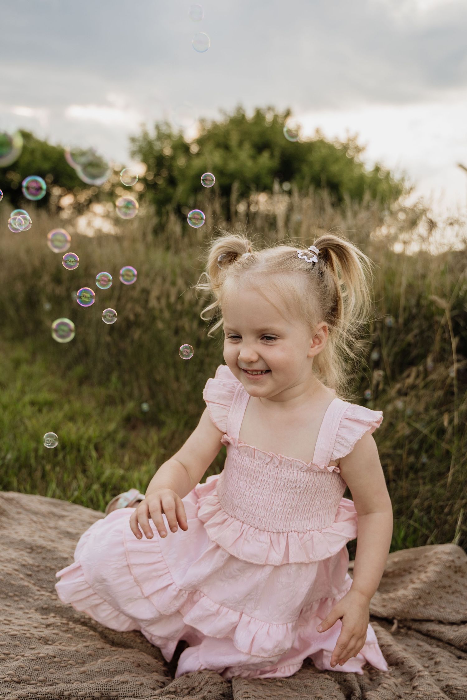 Adorable blonde toddler girl in a pink ruffled dress laughing while playing with soap bubbles outdoors.