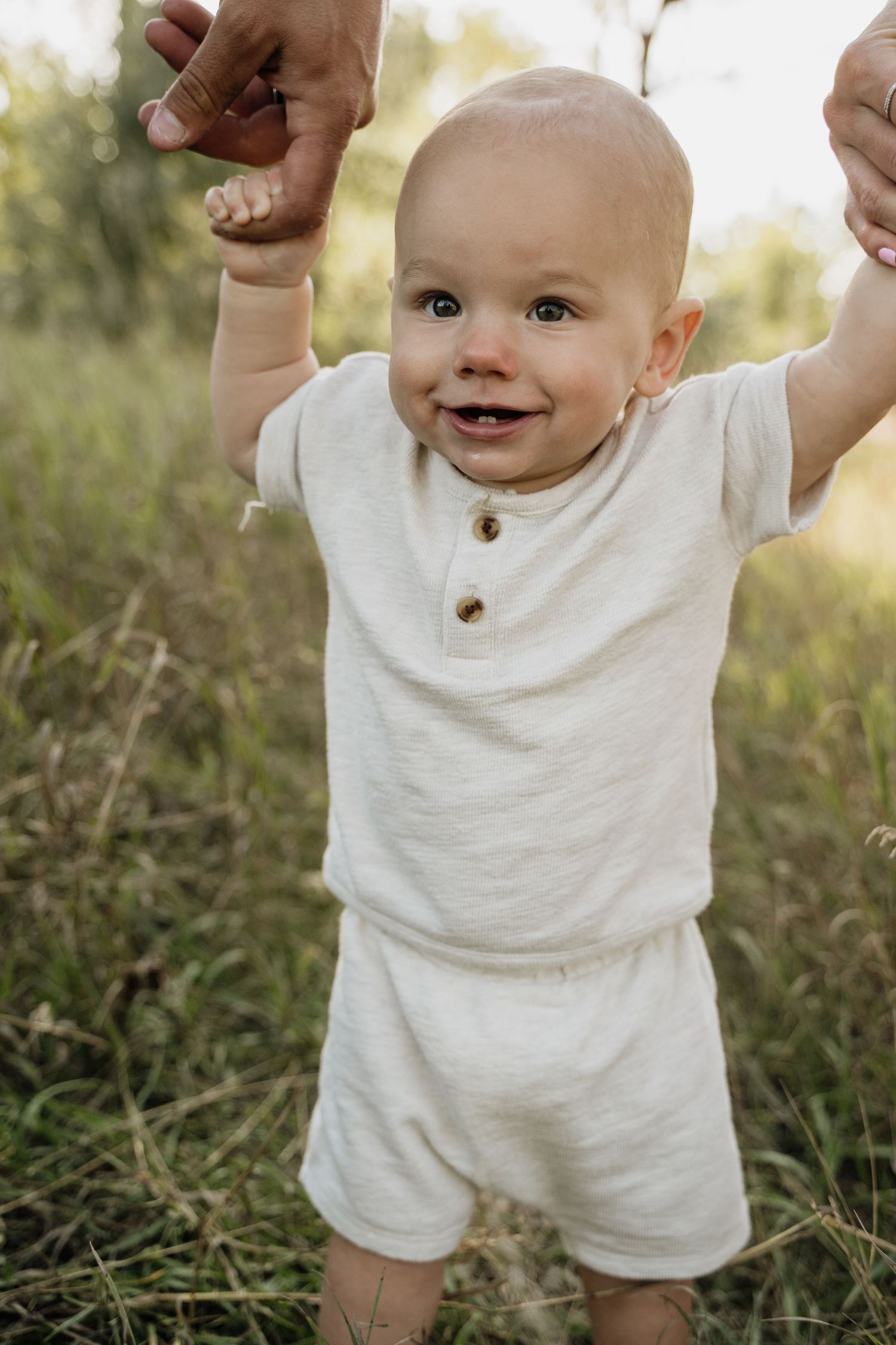 A smiling baby in a white linen outfit stands in a grassy field, supported by an adult holding their hands.
