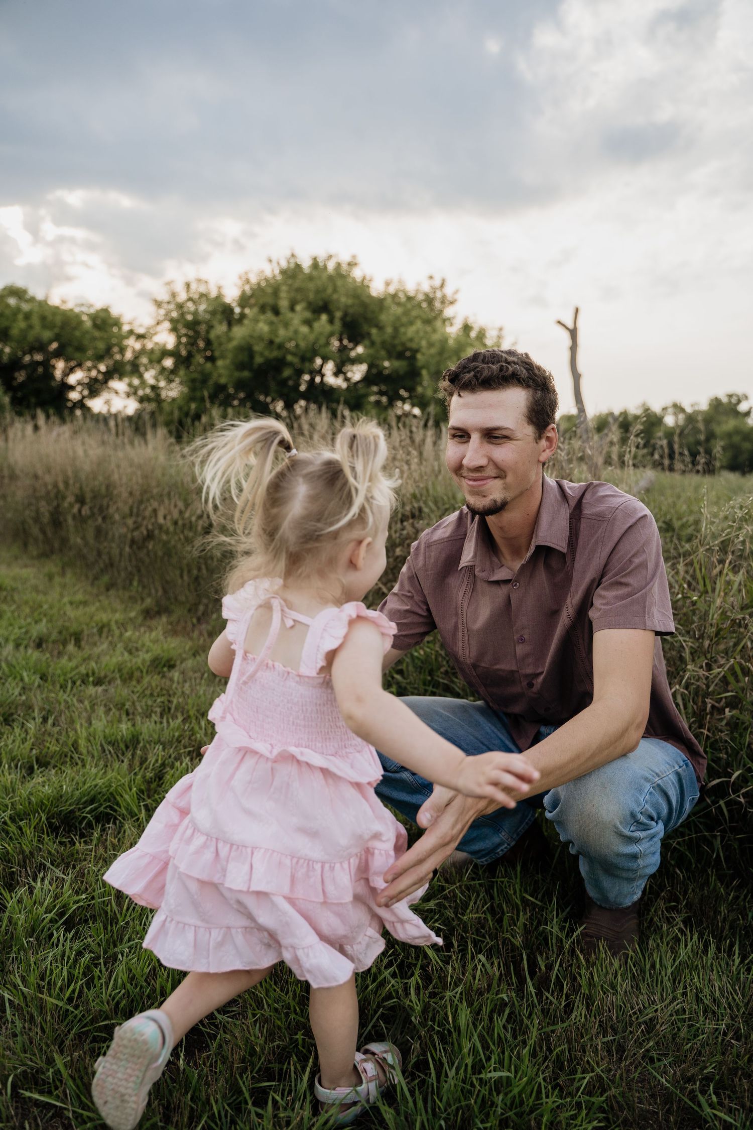 Smiling father kneeling in a grassy field as his young daughter in a pink dress runs toward him.