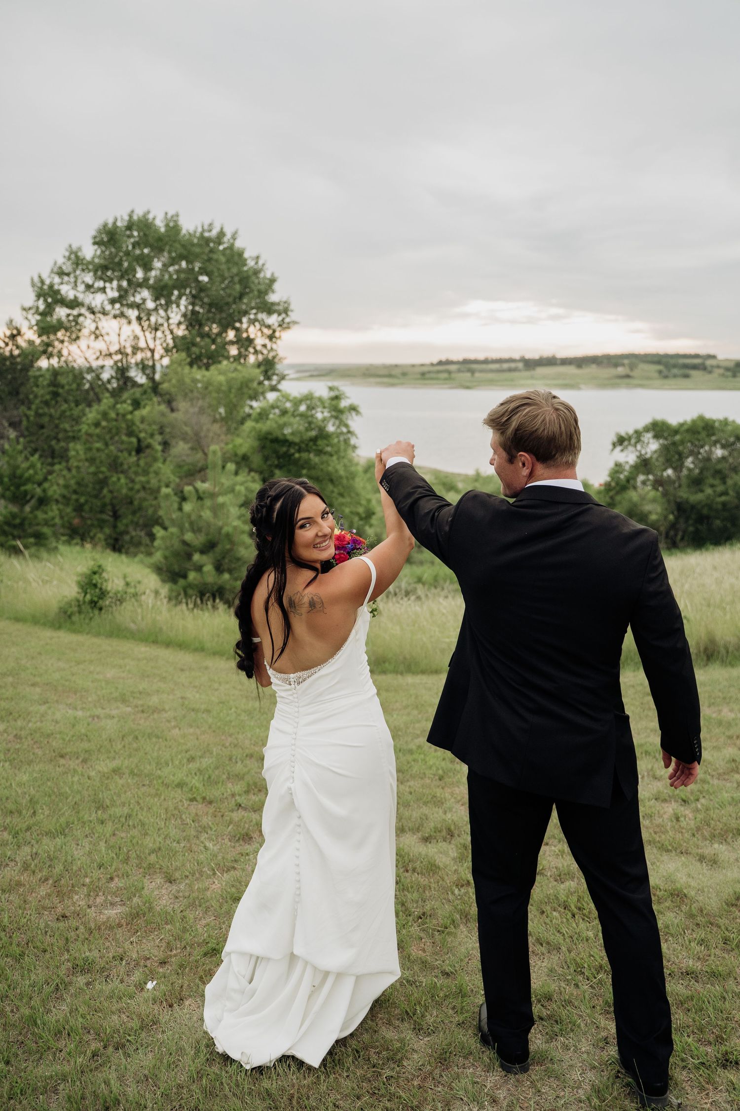 Bride smiling over her shoulder as groom twirls her in a green meadow near a lake.