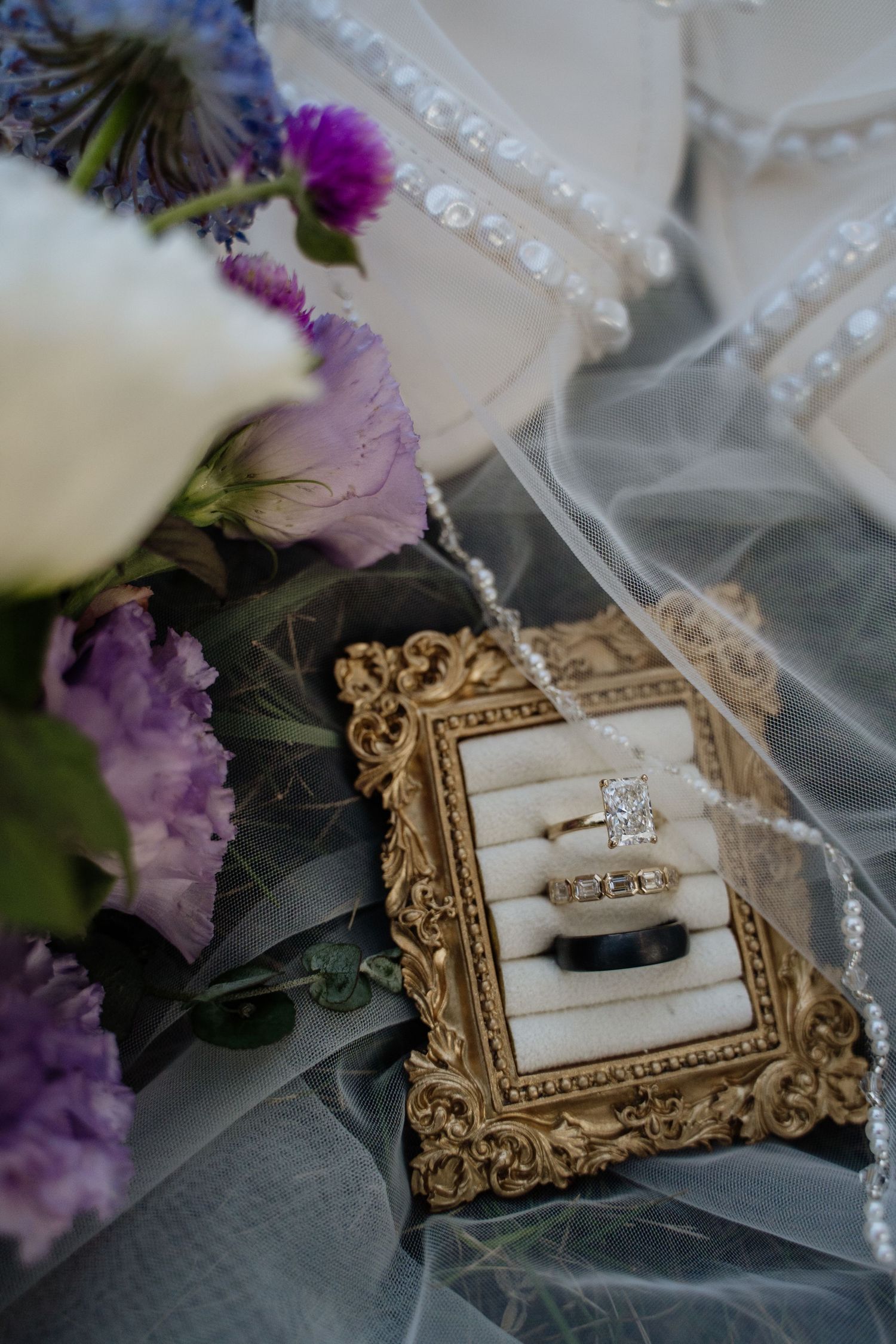 Wedding rings displayed in an ornate gold frame surrounded by purple and white florals and veil.