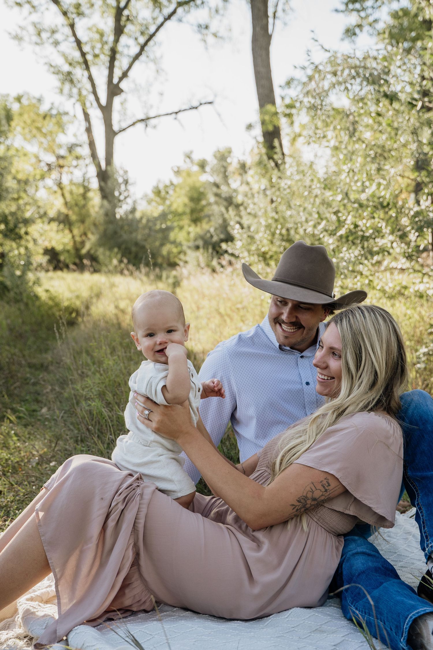 A young family sits on a blanket outdoors, with a man in a cowboy hat and woman in pink holding their baby.