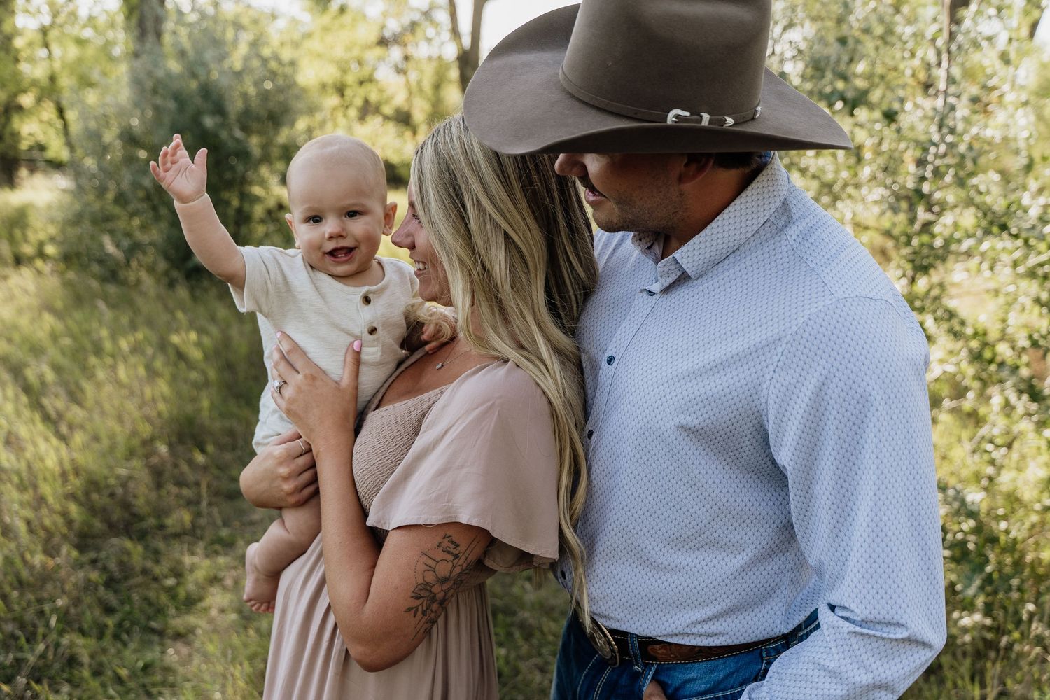 A couple in a green outdoor setting, woman holding a waving baby, man in cowboy hat leaning in close.