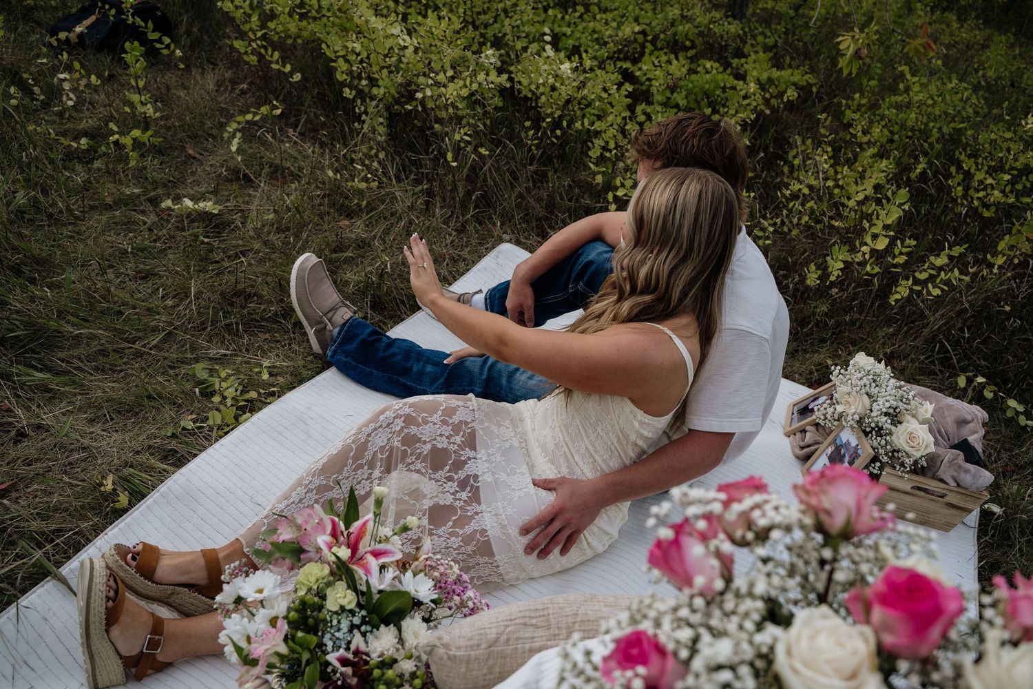 Couple relaxing on a white blanket outdoors surrounded by pink and white floral arrangements in a lush green setting.